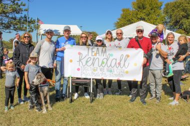 burden of FA | Friedreich's Ataxia news | Kendall Harvey stands behind a placard that reads, "Team Kendall," alongside members of her cycling team at rideATAXIA 2021