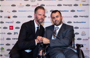 Ethan Ash and Matthew Lafleur, both dressed in suits and ties, pose before a publicity board at the 2022 MM+M Awards event. Matthew is seated in a wheelchair, and to his right, Ethan kneels. They are fist-bumping.