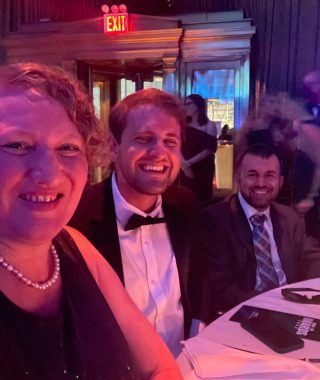 At the BioNews table at the MM+M Awards ceremony, Kellie Benn, Brad Dell, and Matthew Lafleur sit next to one another. All are elegantly dressed, with Kellie wearing a black dress and a white necklace, Brad is in a tuxedo, and Matthew is in a suit and tie. 