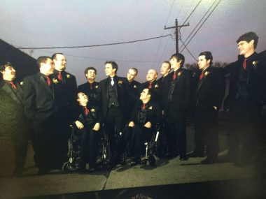 A wedding photo full of a bunch of men, who are the groomsmen. They are all wearing black suits with red ties. The groom stands in the center; two men in wheelchairs are on either side of him, and the other 11 people are standing. It's nighttime and in the background are telephone wires