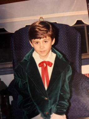 A photo of a young boy in front of a blue chair. He wears a green jacket, white shirt, and a red tie.