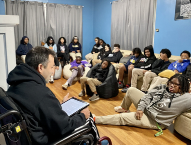 A man in a black hoodie sits in a wheelchair with his back partially to the camera, facing a group of about 18 students sitting on the floor or on a long couch in a blue room with hard wood floors and gray curtains. The man has a tablet on his lap and is speaking to them.