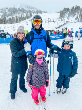 A family of four — including a husband, wife, 8-year-old boy, and 5-year-old girl — post at the base of a ski slope. The man and the two kids are wearing ski helmets and goggles and carrying ski poles. The woman is standing on her husband's right and holding on to his arm.