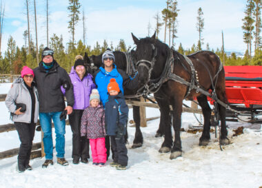 Six people pose next to two large horses, pulling what appears to be a red sleigh. The ground is covered with snow, and they're all bundled up in ski jackets and hats.