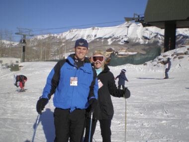 A young couple poses on the slopes near a chairlift in Telluride, Colorado. Both are wearing ski jackets and holding ski poles, and the woman is wearing pink-framed ski goggles.