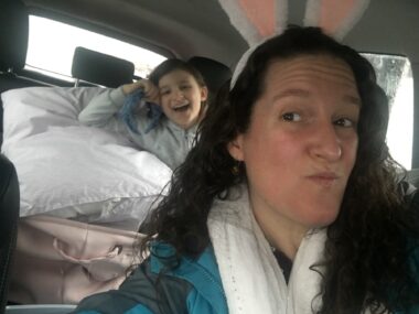 A woman takes a selfie of herself and her young daughter while traveling in the car. In honor of Easter, the woman is wearing bunny ears, as well as a blue jacket and white scarf. Her daughter sits in the back seat, leaning on a large pillow and smiling big.