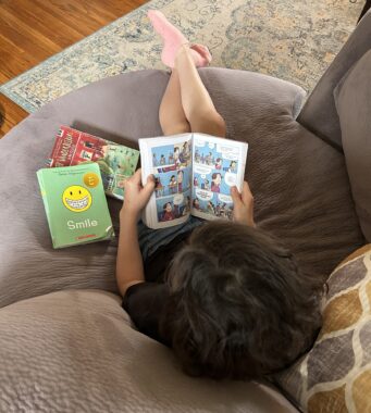 An aerial photo is taken of a young girl sitting on a brown sofa and reading a graphic novel. Her legs are kicked out in front of her, and she has two other books lying to her left.