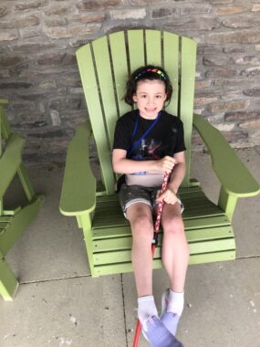 A young girl sits in a lime green Adirondack chair in front of a brick wall at camp. She's wearing a T-shirt, shorts, and purple shoes and is holding her red walking stick between her legs.