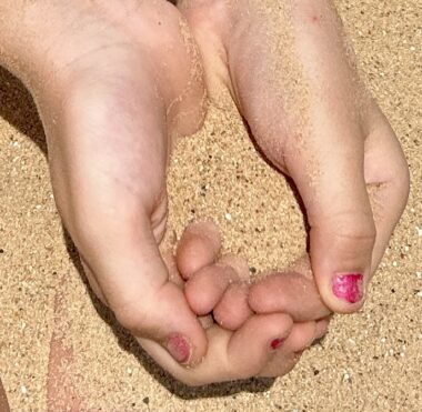 A closeup of an 11-year-old girl's cupped hands holding sand on the beach. Her fingernails are painted pink and a bright sun reflects off each grain of sand. 