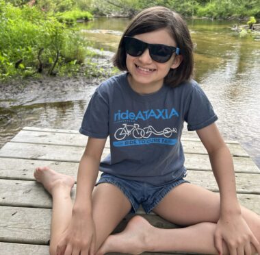 A young girl in denim shorts, black sunglasses, and a blue "rideATAXIA" T-shirt sits on a dock. A river, lake, or pond seems to be behind her, with low, free-form greenery around it.