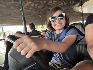 A low-angle photo shows a girl driving a bumper car and grinning from ear to ear. She has shoulder-length brown hair and is wearing a grayish blue T-shirt and sunglasses with white flowers in the corners. The photo appears to be taken by the girl's mom, who is just visible in the seat next to her.
