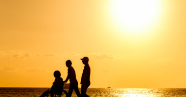 Two people walking with a person in a wheelchair at sunset on the beach