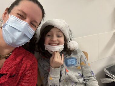 A woman takes a selfie with her 10-year-old daughter at the hospital. The girl is wearing a gray sweater, a silver Santa hat, and a white face mask, which she's pulled down to her chin to smile for the photo. Her mom is wearing a red sweater, and a blue mask covers the bottom half of her face. They're sitting in chairs against a white tiled wall.
