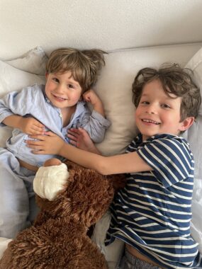 Two boys are seen from overhead as they lie on a bed, with a brown stuffed animal between them. The younger boy on the left has golden brown hair and wears light blue pajamas; the boy on the right has brown hair and wears a blue-and-white-striped shirt. He is reaching over to put his hands on the younger boy's midsection.