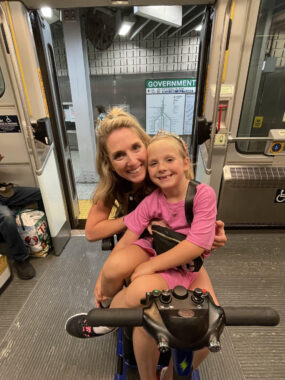 A blond woman sits on a mobility scooter with her daughter, blond and in pink, in her lap. They are on the subway, with the doors behind them still open to the platform.