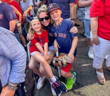 A mom in her 30s sits in a chair surrounded by a crowd of people waiting in line. She has her arms wrapped around her son, who's standing next to her, and her daughter, who's sitting on her lap. They're decked out in red and blue for a Red Sox game.