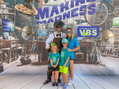 A family of four poses for a photo in front of a backdrop that looks like a scientific laboratory. The words "Making Madness" are featured in large purple text, and a computer screen reads "Lake Hill Church VBS."