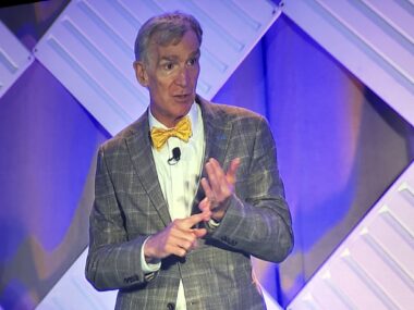 "The Science Guy," Bill Nye, stands on a stage speaking to a crowd. He's wearing a signature yellow bow tie and a plaid suit. He's pointing to his left hand with his right hand to emphasize a point.