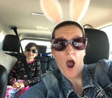 A woman wearing a denim jacket, sunglasses, and peach and white rabbit ears sits in a car's driver's seat. Her mouth is rounded as if saying "Oh!" A young girl wearing sunglasses and a black top with mostly pink designs sits in the back seat. 