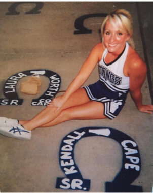 A young woman wearing a white and blue cheerleading uniform sits on the ground next to a horseshoe with her name painted on it.