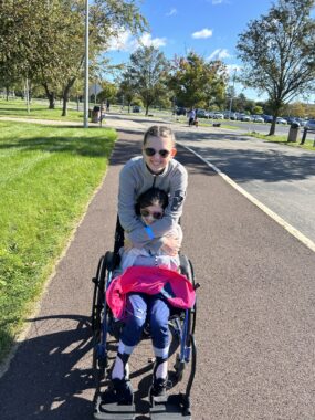 Two young sisters, one 11 and the other a few years older, pose together for a photo on what looks like a bike trail along a park. The younger girl is wrapped in a pink vest in a wheelchair and is embraced by the older girl, who stands behind her. Both are wearing sunglasses, as it's a sunny, bright, and cloudless day. There are trees in the background and cars parked in a parking lot. Both girls are smiling broadly.