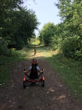 On a wide dirt path between two stands of trees and other greenery a young girl rides a trike. A woman is far ahead of her on the path.