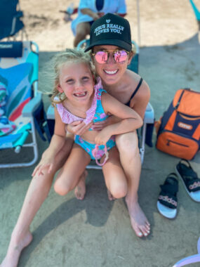 A woman sits on a beach chair and holds her 7-year-old daughter on her lap. Both are smiling and wearing swimsuits, and the mom has on a black baseball cap and sunglasses. There are several other chairs set up on the sand around them, and they appear to be sitting under some sort of canopy.