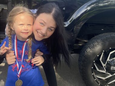 A young woman with long dark hair leans down to hug a young girl and smile together for a photo. The little girl has blond braids and is wearing a medal over a blue T-shirt with a red graphic and writing. The words 