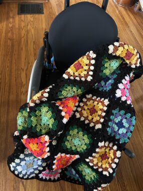 A granny square blanket sits on a chair in a wooden-floored room of a house. Colorful blue, yellow, and orange squares are set off by black borders.