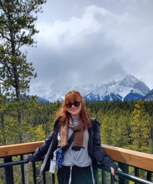 A woman with reddish-brown hair, sunglasses, gray shirt, brown scarf, and a blue jacket stands at a fence corner. Behind her are acres of green trees and the peaks of bluish, snow-covered mountains, rising into the overcast clouds.