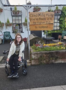 A woman with long, reddish-brown hair, glasses, a white sweater, and dark shirt, pants, and shoes sits in a wheelchair outside a white building. To her side is a flower bed with a sign above it that reads "You've Made it to the Highest Pub In Ireland."