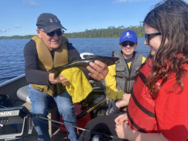 Three people sit in a boat on a lake. All of them are wearing life jackets. A man on the left, wearing a worn baseball cap and sunglasses, smiles as he displays a large fish to a teenage girl who studies it proudly. Another woman looks on.