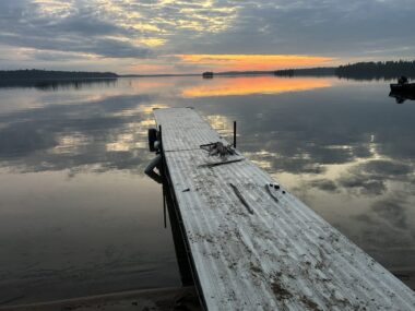 The sun rises over a small lake, while an old dock stretches out in the middle of the frame. Blue-gray skies, with a hint of reddish orange, are reflected in the water.