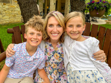 A woman sits on a wooden bench between her two children, one boy and one girl. They are dressed nicely in pastel colors, and all are smiling happily.