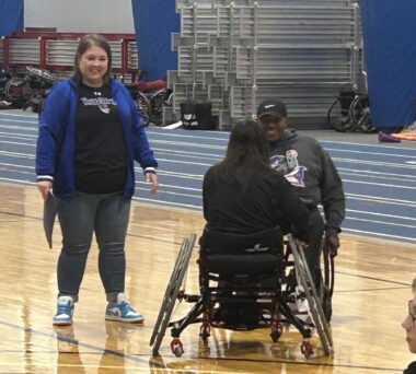 Two basketball coaches – one standing and one using a wheelchair – chat with one of their players on the court, a teenage girl using a wheelchair.