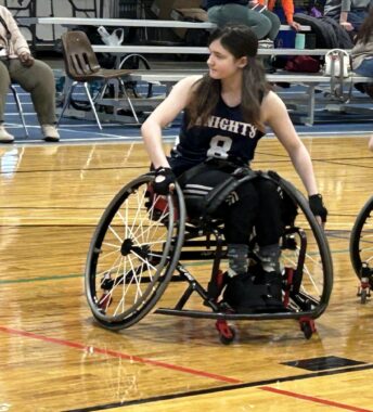 A teenage girl wearing a black jersey navigates a basketball court in a wheelchair.